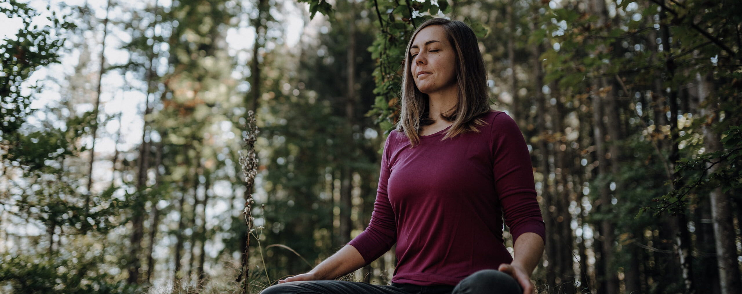 Franziska sitzt entspannt im Schneidersitz auf einem Felsen im Wald und hat die Augen geschlossen.