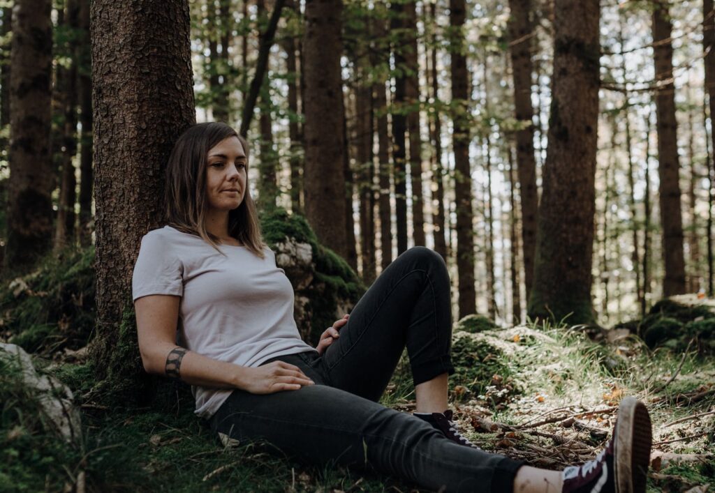 Franziska sitzt an einen Baum gelehnt in einem Wald, wo die Sonne reinscheint. Sie hat ein Bein angewinkelt und eines ausgestreckt.